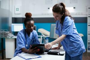 diverso médico cuidado de la salud personal equipo mirando a digital tableta pantalla en moderno profesional clínica oficina. africano americano enfermero comparando datos con compañero de trabajo en hospital lugar de trabajo foto