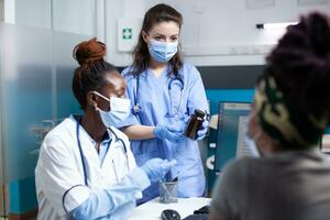 Nurse and doctor offering patient medical guidance in clinic office during appointment. General practitioner in antibacterial protective mask and gloves prescribing medicine in bottle to woman photo