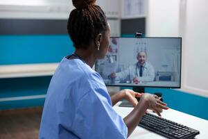 africano americano enfermero a escritorio en un vídeo reunión comunicado con médico compañero de trabajo en un moderno médico oficina. general facultativo en telesalud videollamada con colega foto