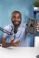 Smiling vlogger presenting camera battery, reviewing videography equipment while recording video. Blogger promoting photography gear while live streaming on social media channel photo