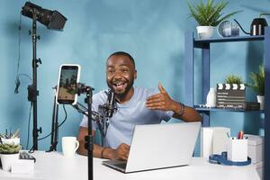 African american blogger speaking and using laptop while recording video on smartphone for internet channel. Online streamer broadcasting live using mobile phone on tripod photo