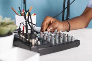 hombre mano torneado mando en sonido controlador equipo en estudio cerca arriba. africano americano blogger utilizando música mezclador consola profesional engranaje mientras creando digital contenido para vlog foto