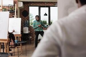 Young African American man teaching art to people in creative studio, male instructor demonstrating sketching techniques to students sitting at easels during group drawing workshop photo