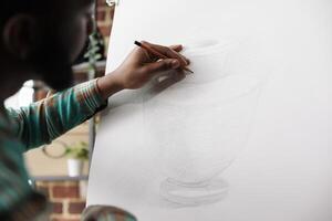 Student guy drawing with pencil on canvas. African American man artist staying focused while creating art in studio or workshop, sitting at easel sketching basic vase using pencil photo