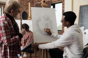 Young African American guy instructor teaching senior woman to draw during art class, teacher sitting at easel showing mature student how to hold and use pencil. Taking up drawing lesson in retirement photo