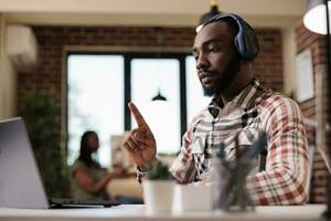 africano americano estudiante con inalámbrico auriculares en en línea vídeo conferencia lección señalando arriba haciendo mano gesto en frente de ordenador portátil. masculino programador en intenet llamada levantamiento índice dedo. foto