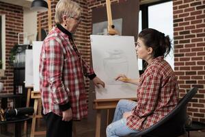 Friendly smiling senior woman teacher standing near easel working with student during drawing lesson. Adult daughter attending group art class with mother, spend time together while learning to draw photo