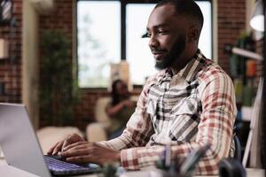 retrato de empresario trabajando remoto mecanografía y mirando a ordenador portátil pantalla mientras esposa es acecho televisión desde sofá. africano americano persona de libre dedicación utilizando portátil computadora mientras roomate es relajante. foto