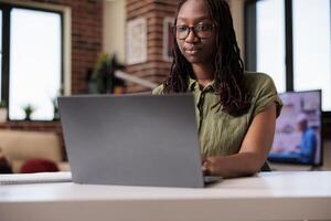 Portrait of confident freelancer looking at laptop screen and typing while working from home on business project. African american employee doing remote work on portable computer at desk in living photo