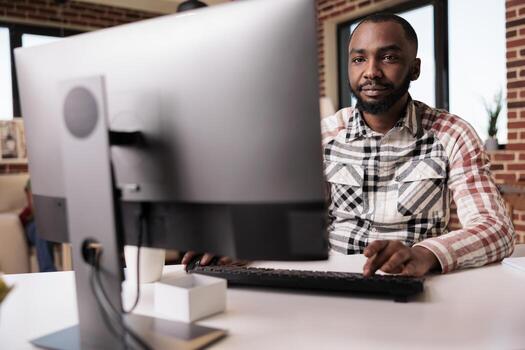 Confident african american programmer looking at computer screen and typing while working remote from home living room. Selective focus on freelancer using pc for remote job sitting at desk. photo