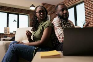 Upset african american girlfriend threatening man while arguing about relationship sitting back to back on couch. Couple living together having a fight about marriage problems in home living room. photo