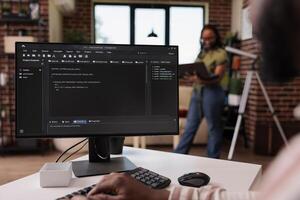 Freelance programmer developing source code on personal computer while girlfriend is using laptop standing in living room. African american couple working remote programming software together. photo