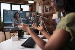 africano americano mujer en casual reunión con puesta en marcha propietario y equipo en vídeo llamada desde hogar vivo habitación. trabajo buscador en en línea entrevista en personal computadora mientras compañero de habitación es relajante leyendo un libro. foto