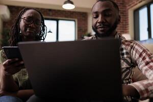 Woman browsing social media on smartphone while boyfriend is watching online series on laptop while sitting on couch. African american couple spending free time together looking at video content. photo