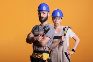 Serious construction workers posing with pair of pliers and hammer, holding slegdehammer and renovating tools in studio. Man and woman working as constructors with overalls and helmet. photo