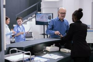 Elderly patient standing at hospital reception counter paying medical consultation with phone using contactless, talking with receptionist in lobby. Old man making payment after having examination photo