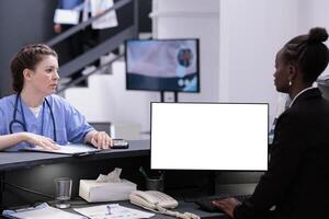 African american receptionist using pc with white isolated screen at reception counter desk in hospital. Diverse medical staff working with mockup template during checkup visit consultation photo