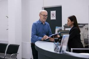 Elderly patient discussing disease expertise with asian receptionist while signing medical paper, standing at hospital counter in hospital waiting area. Health care service and concept photo