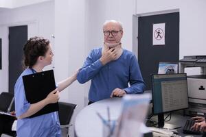 Elderly patient with medical neck collar having checkup visit examination with physician assistant in hospital lobby. Man with cervical foam and nurse discussing about symptoms and medication photo