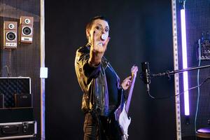 Brunette guitarist with short hair doing rockstar gesture at camera while playing electric guitar in studio, preparing to perform rock music at metal concert. Performer blowing bubble gum photo