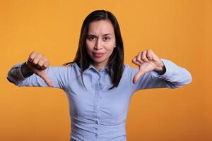 Unhappy asian model giving thumbs down negative sign in studio, doing disapproval and bad symbol. Disaproval woman expressing disagreement with dislike gesture over yellow background photo