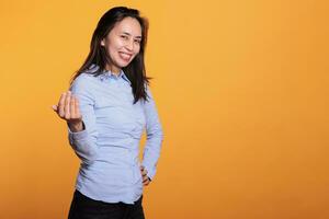 Filipino model inviting you to come here with a friendly smile on her confident expression, posing in studio over yellow background. Positive brunette woman is calling out with a finger gesture photo