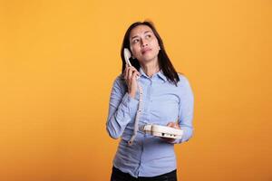 Filipino woman talking at landline phone with remote friend, chatting on retro telephone line with cord in studio over yellow background. Cheerful young adult enjoying discussin during break time photo