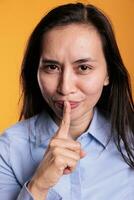 Cheerful serious filipino woman making silence gesture by putting forefinger over lips posing in studio over yellow background. Young adult doing shh symbol, expressing confidentiality photo
