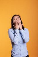 Overworked sleepy asian woman covering mouth with hand being tired while posing in studio standing over yellow background. Lazy adult yawning having exhausted expression, working overtime photo