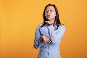 Confident joyful filipino woman blowing air kissed during studio shot, posing over yellow background. Attractive model expressing love, looking at the camera with a flirty smile. Romantic gesture photo