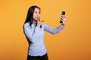 asiático mujer haciendo shh gesto durante videollamada reunión, teniendo secreto discusión con remoto amigo en estudio terminado amarillo antecedentes. alegre adulto disfrutando remoto conversacion durante descanso foto