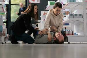 Frightened women rescuing man suffering from epilepsy attack, holding unconscious person and waiting for help in drugstore. Pharmacy asian and caucasian clients noticing epileptic seizure accident photo