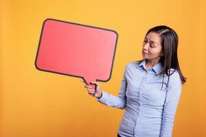 Asian young adult holding empty red speech bubble with copy space, advertising beauty product. Woman standing in studio over yellow background, looking at blank dialog frame with place for text photo