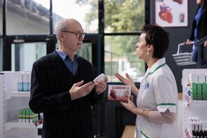 Chemist discussing with elderly customer heart disease prevention and recommending supplements. Old man explaining cardiovascular symptoms to pharmaceutical assistant, choosing medication photo