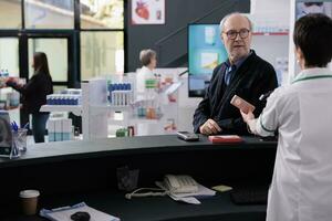 Pharmacy elderly customer watching cashier scanning purchase at paydesk, buying sunscreen and heart supplement in drugstore. Senior man in glasses standing at counter desk and waiting to pay photo