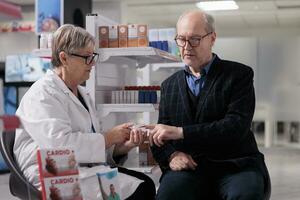 Chemist using cardiology device for old patient oxygen saturation measurement in drugstore. Senior man in glasses making pulse oximetry test and getting cardiovascular disease diagnosis photo