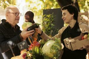 Young friendly female farmer selling organic veggies from produce stand at farm market talking with customer. Local vendor showing eggplant to senior man buying fresh and healthy locally grown food photo