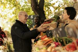 Senior man buying various natural farm products at farmers market stand, vendor giving box of organic fresh products. Old person holding colorful farming eco fruits and veggies. photo