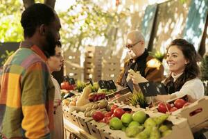 Young multiracial couple man and woman buying fresh organic produce at farmers market. Smiling local female vendor standing behind fruit and vegetable stand offering customers to try apple photo