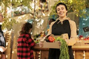 Young cheerful seller fruits and veggies market stand owner showing fresh organic produce box and smiling at camera. Female farmer selling healthy local food from roadside stand, bio products. photo