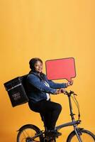 Smiling african american restaurant employee holding red speech bubble promoting takeaway food service while delivering fast food order to client using bike as transportation. Takeout concept photo