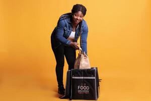 Takeout delivery worker taking out paper bag from thermal backpack delivering takeaway food order to client during lunch time. Deliverywoman standing in studio with yellow background photo