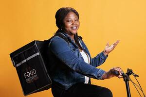 Pizzeria courier with takeout thermal backpack showing takeaway advertisement while riding bike in studio with yellow background. Employee delivering fast food order to clients during lunch time photo