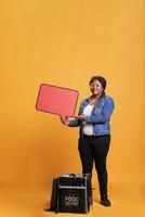 Portrait of deliverywoman holding red speech bubble advertisting takeaway food service while standing in studio with yellow background. Restauran courier carrying thermal backpack photo