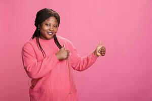 Cheerful young adult doing thumbs up gesture in front of camera doing approval and agreement gesture with finger, smiling in studio. Pleased woman showing okay and positive sign . photo