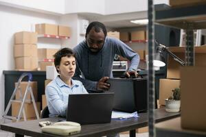 Diverse team talking about metallic box while checking transportation logistics for package delivery on laptop computer. Storehouse workers analyzing online shipping order working in warehouse photo