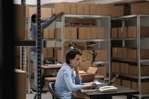 Storehouse supervisor looking at cardboard box checking package shipping details before preparing delivery, typing shipping detalies on laptop. Worker analyzing cargo stock working in warehouse photo