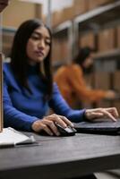 Asian warehouse operator coordinating order shipping on laptop. Storehouse young woman employee managing freight distribution and supply chain operations on computer in storage room photo