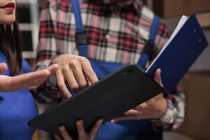 Warehouse operators checking inventory data on digital tablet and analyzing orders checklist on clipboard. Storehouse postal managers colleagues comparing quality control reports photo