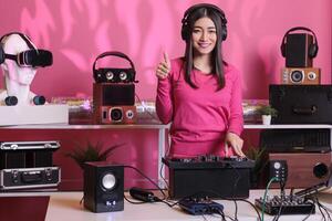 Positive musician standing at dj table doing thumbs up gesture while mixing electronic music and techno using mixer console. Asian artist playing stereo sounds with electronics and microphone photo